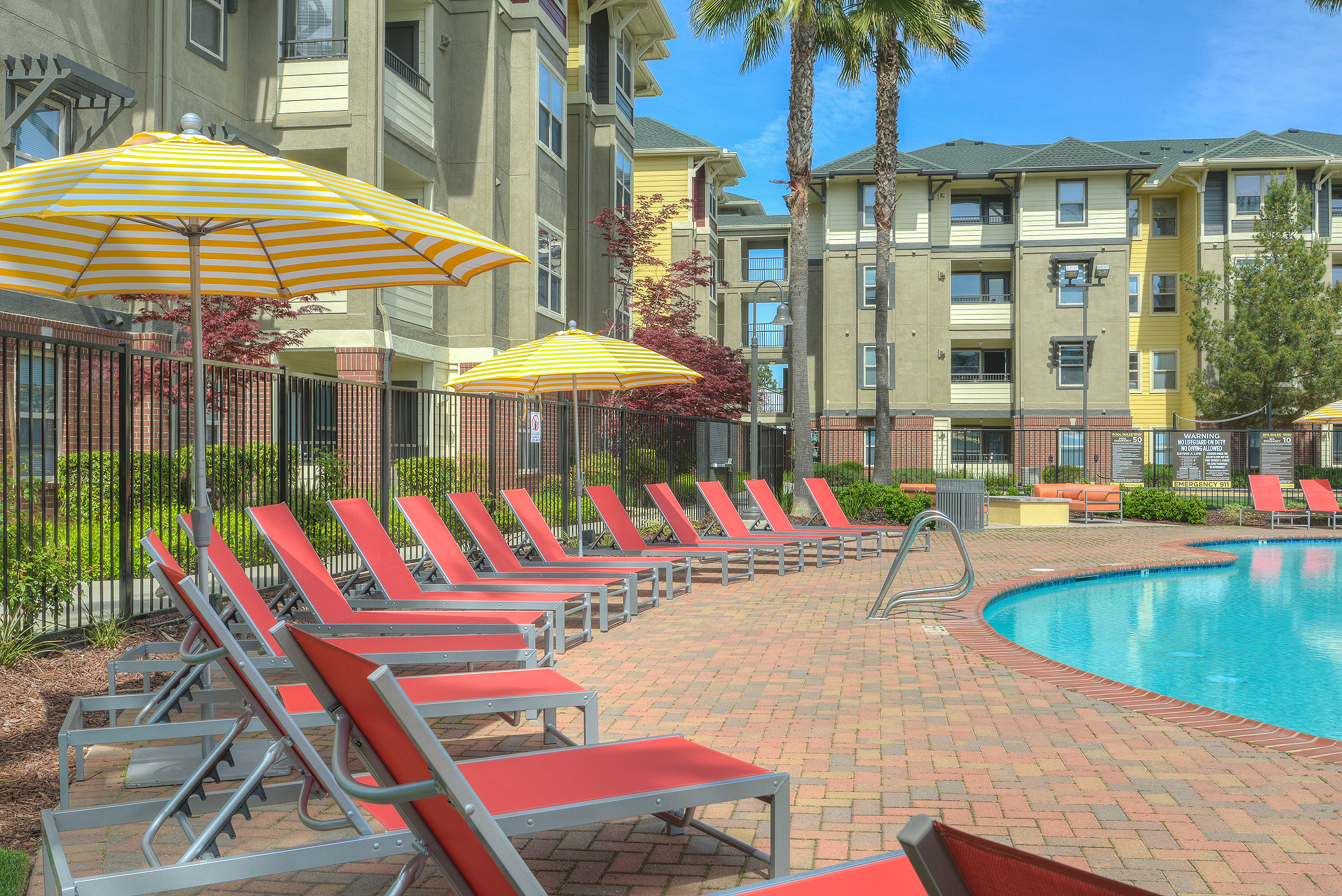 Exterior of pool with brick pool deck rimmed with lounge chairs with 4-story apartment buildings in background