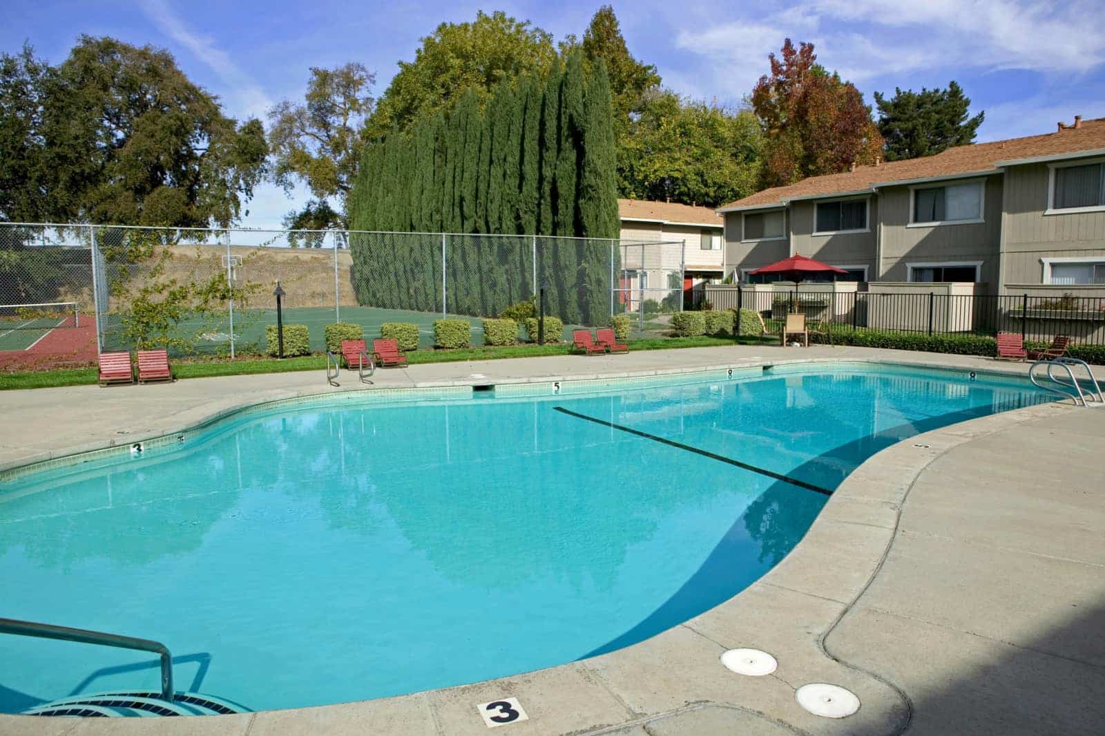 View of the pool with a lawn, tennis court and apartment buildings in the background.